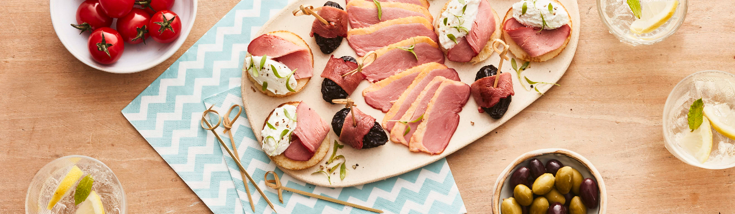 Planche apéro composée de lamelles de fumés séchés de canard, de blinis au chèvre et fumé séchés et de roulés pruneaux et canard fumé