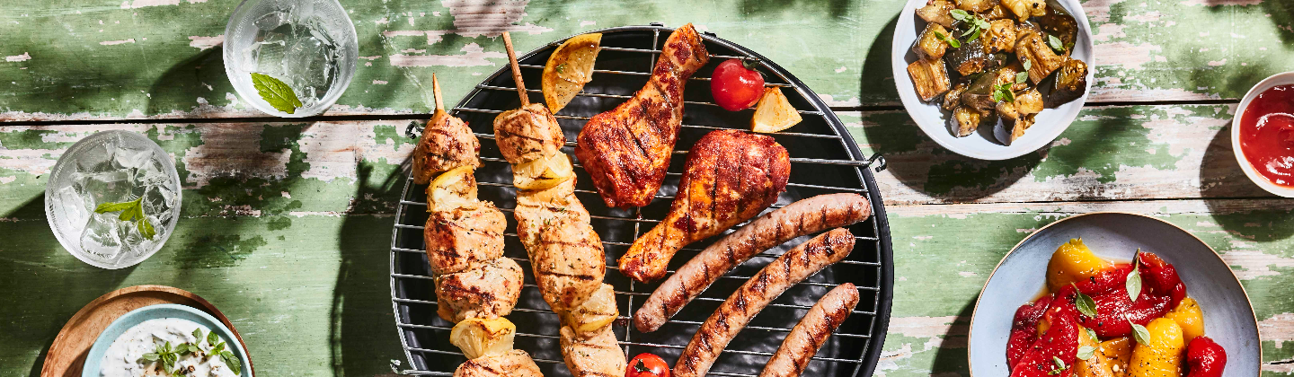 Assortiment de brochettes thym-citron, saucisses et pilons paprika sur une grille de barbecue accompagnés de poivrons et courgettes grillés